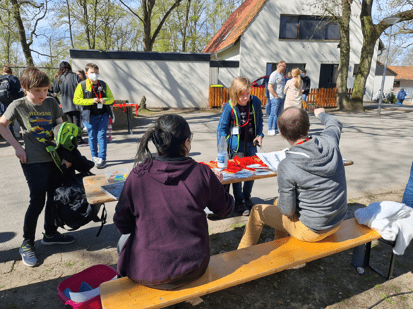 Gruppendiskussion im Freien mit Kindern und Erwachsenen an einem Tisch, umgeben von B&auml;umen und Geb&auml;uden.