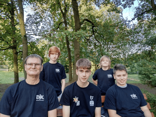 F&uuml;nf Personen in schwarzen T-Shirts sitzen auf einer Bank im Park, umgeben von B&auml;umen und nat&uuml;rlichem Licht.