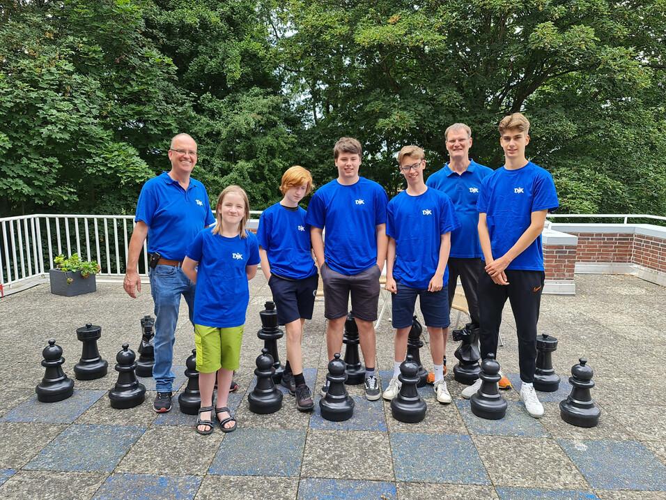 Gruppe von sieben Personen in blauen T-Shirts steht l&auml;chelnd vor gro&szlig;en Schachfiguren auf einem Au&szlig;enbereich.
