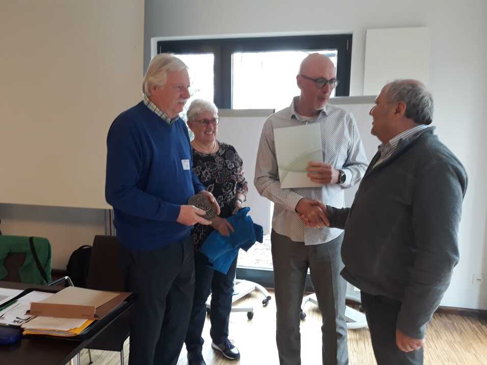 Vier Personen stehen in einem Raum und unterhalten sich, w&auml;hrend eine Person eine Urkunde &uuml;bergibt.