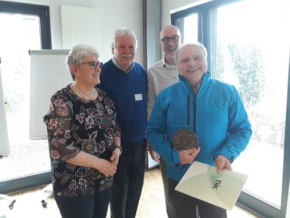 Vier Personen stehen in einem hellen Raum, eine Person h&auml;lt eine Medaille und ein Zertifikat in der Hand.