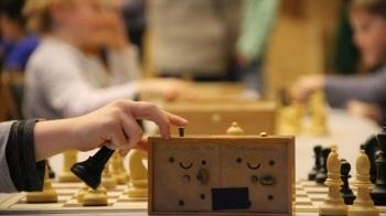 Nahaufnahme einer Hand, die eine Schachfigur hebt, mit Schachbrett und weiteren Spielern im Hintergrund.