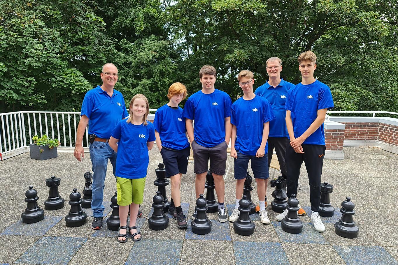 Gruppe von sieben Personen in blauen T-Shirts steht l&auml;chelnd vor gro&szlig;en Schachfiguren auf einem Au&szlig;enbereich.