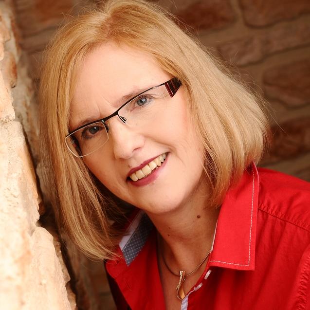 Porträt einer lächelnden Frau mit blonden Haaren in roter Bluse, die an einer steinernen Wand lehnt.