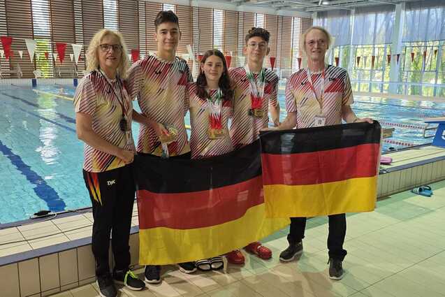 F&uuml;nf Personen in Badekleidung halten eine deutsche Flagge vor einem Schwimmbecken mit Farben von Schwimmbahnen.