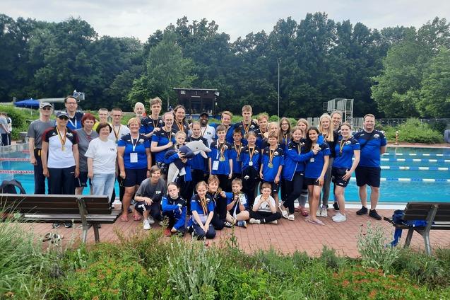 Gruppe von Schwimmern in blauen Shirts mit Medaillen, posiert am Schwimmbeckenrand umgeben von B&auml;umen.