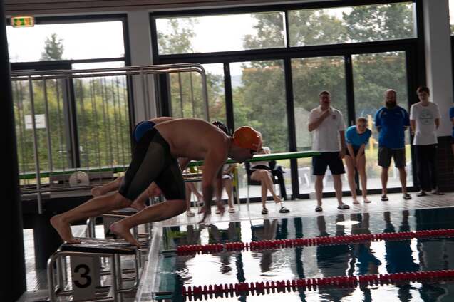 Schwimmer springt vom Startblock ins Wasser, Zuschauer beobachten am Beckenrand in einem Hallenbad.