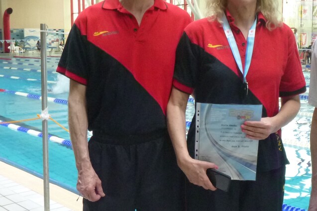 Zwei Schwimmtrainer in roten und schwarzen Trainingsanz&uuml;gen stehen am Beckenrand eines Schwimmbades und halten Unterlagen.