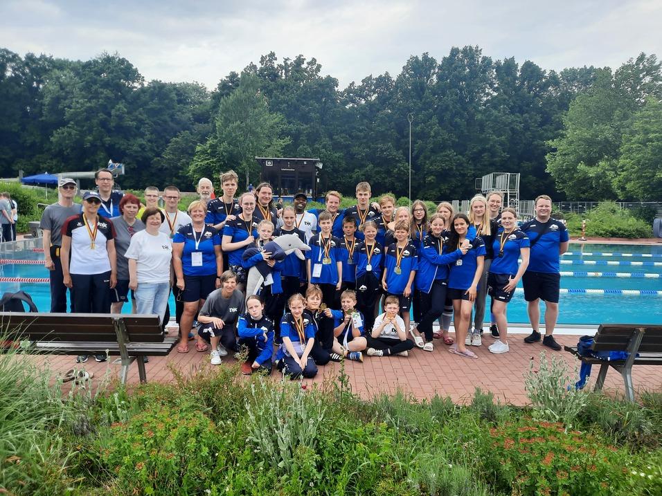 Gruppe von Schwimmern in blauen Shirts mit Medaillen, posiert am Schwimmbeckenrand umgeben von B&auml;umen.