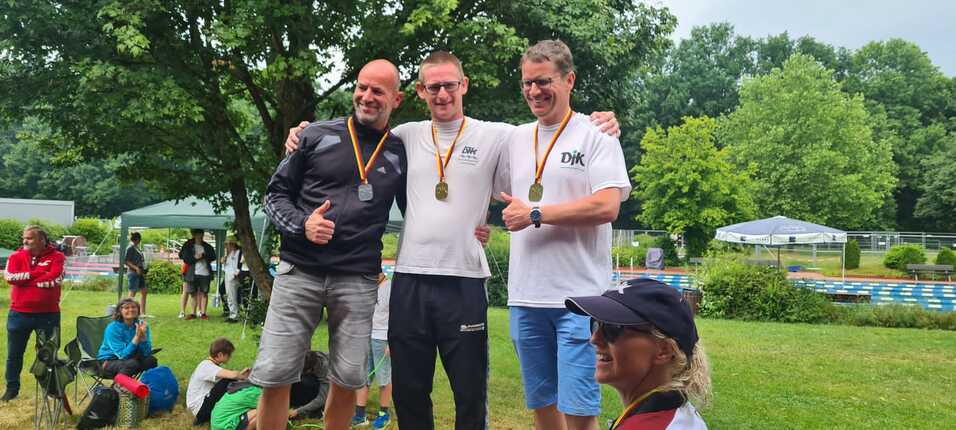 Drei M&auml;nner mit Medaillen posieren l&auml;chelnd in einem Park, umgeben von gr&uuml;nen B&auml;umen und Zuschauern.