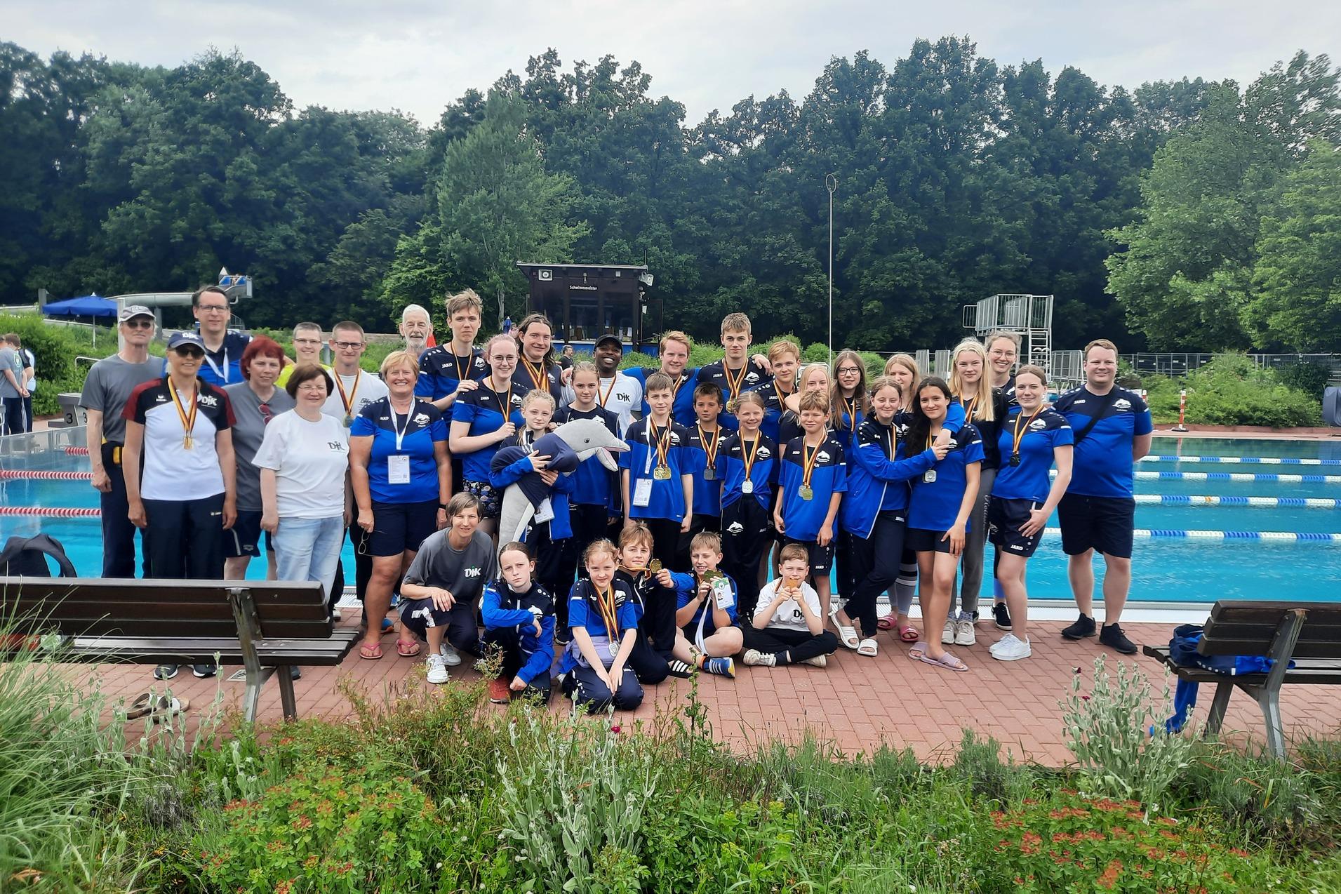 Gruppe von Schwimmern in blauen Shirts mit Medaillen, posiert am Schwimmbeckenrand umgeben von B&auml;umen.