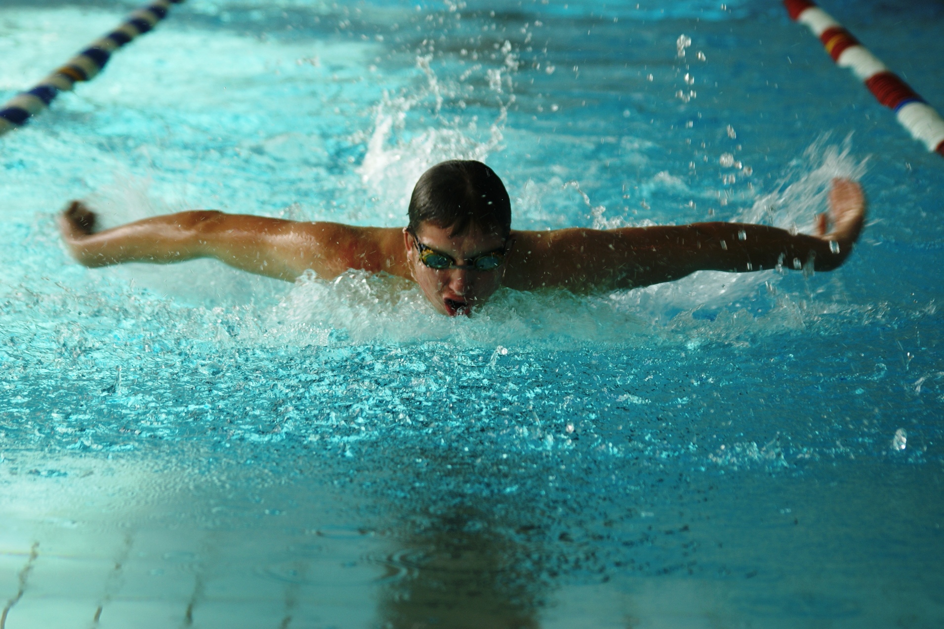 Ein Schwimmer vollzieht den Schmetterlingsstil in einem Hallenbad. Spritzwasser umgibt ihn.