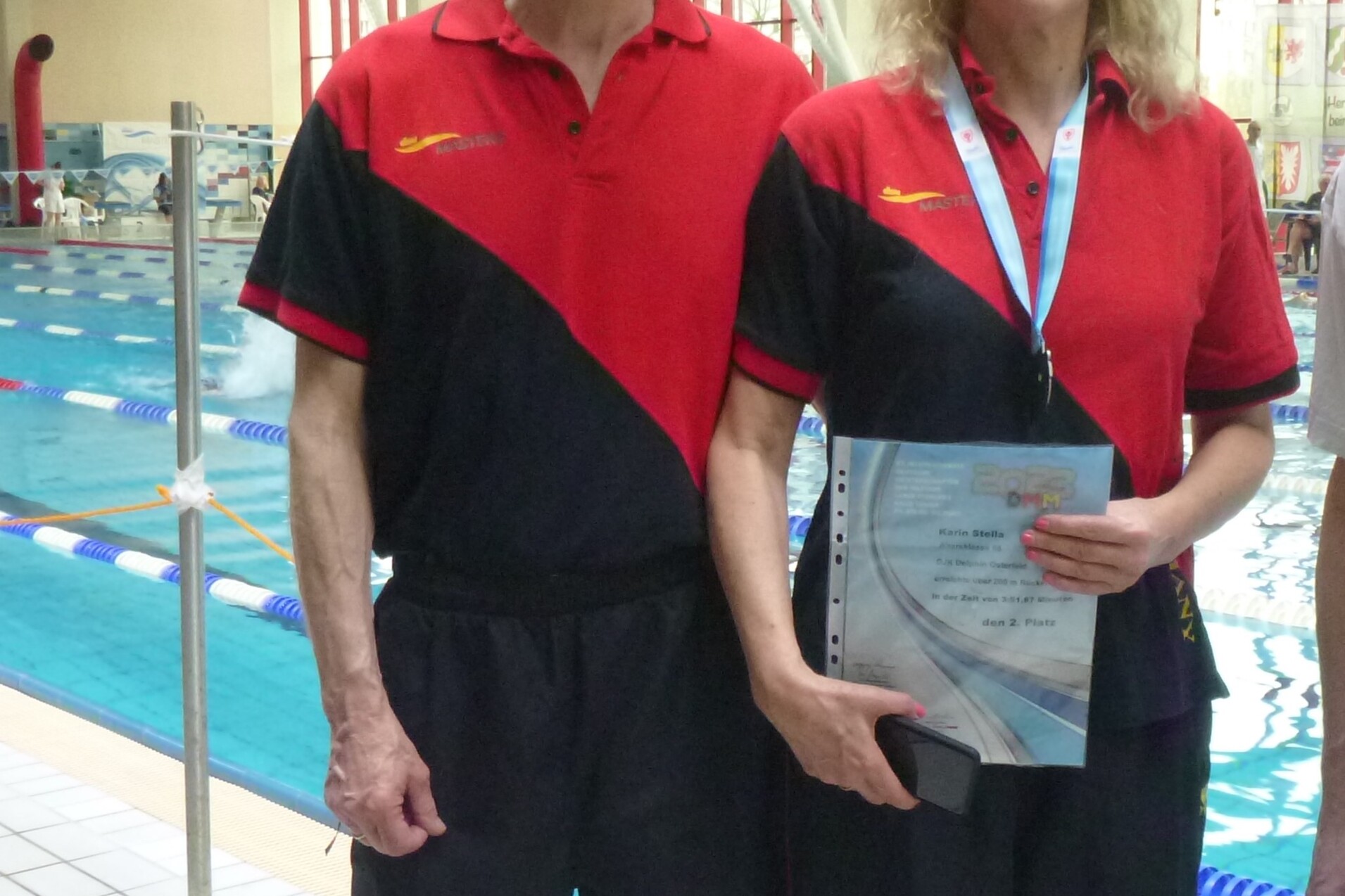 Zwei Schwimmtrainer in roten und schwarzen Trainingsanz&uuml;gen stehen am Beckenrand eines Schwimmbades und halten Unterlagen.