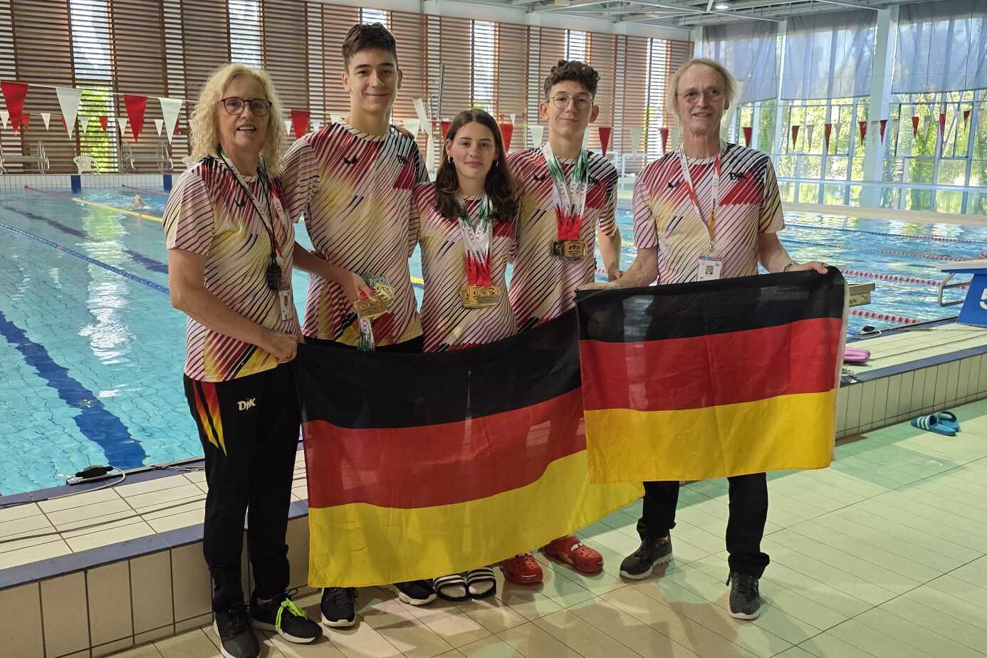 F&uuml;nf Personen in Badekleidung halten eine deutsche Flagge vor einem Schwimmbecken mit Farben von Schwimmbahnen.