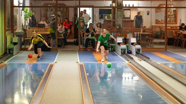 Zwei Spielerinnen in Sportkleidung werfen Bowlingkugeln auf eine Bahn; Zuschauer im Hintergrund beobachten das Spiel.