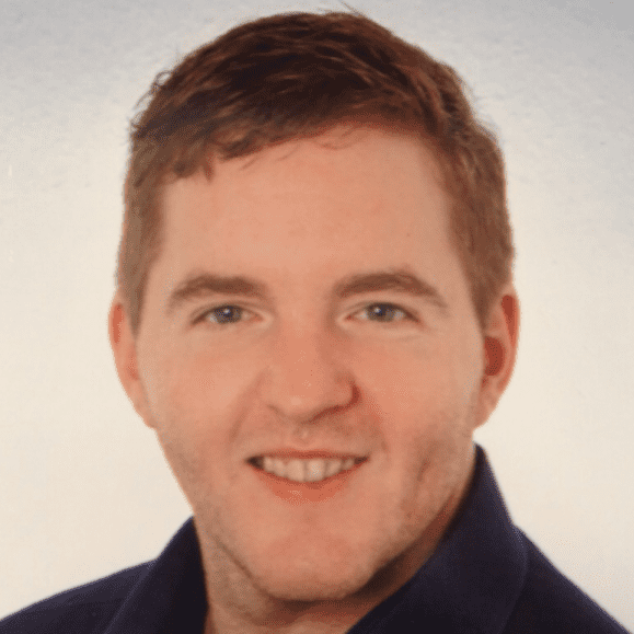 Portr&auml;t eines Mannes mit kurzem, braunem Haar, l&auml;chelnd, in einem dunklen Hemd vor neutralem Hintergrund.