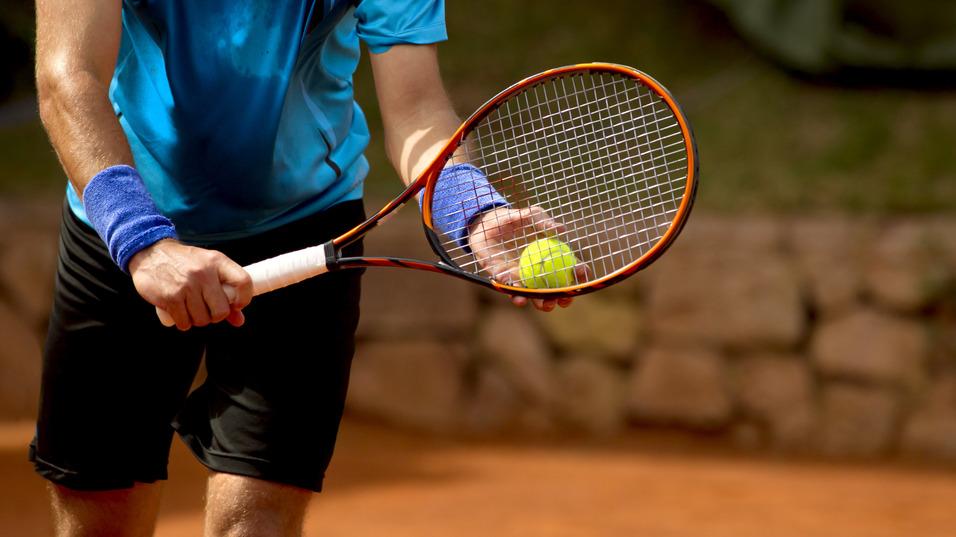 Tennisspieler hält einen Schläger in der rechten Hand und eine gelbe Ball in der linken Hand auf einem Sandplatz.