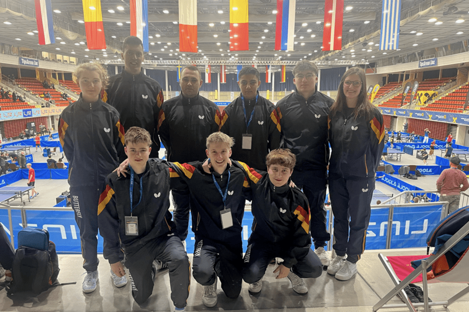 Gruppe von Tischtennisspielern in einheitlicher Sportkleidung in einer Halle mit Flaggen und Tischtennispl&auml;tzen im Hintergrund.