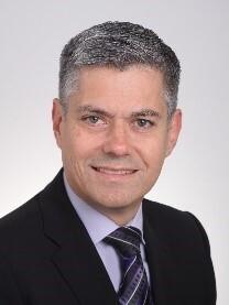 Porträt eines Mannes mit kurzen grauen Haaren, Anzug und Krawatte, lächelt vor hellem Hintergrund.