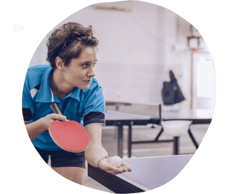 Junge Sportlerin in blauer Kleidung h&auml;lt einen Tischtennisschl&auml;ger und einen Ball vor einem Tischtennistisch.