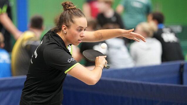 Junge Tischtennisspielerin schl&auml;gt mit einem Schl&auml;ger einen Ball auf einem Tisch, Publikum im Hintergrund.