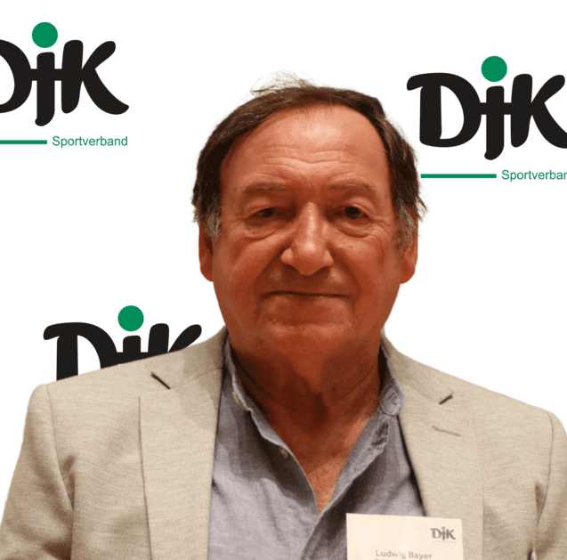 Portr&auml;t eines &auml;lteren Mannes mit braunen Haaren, der ein Sakko tr&auml;gt, vor einem Hintergrund mit dem Logo des DJK Sportverbands.