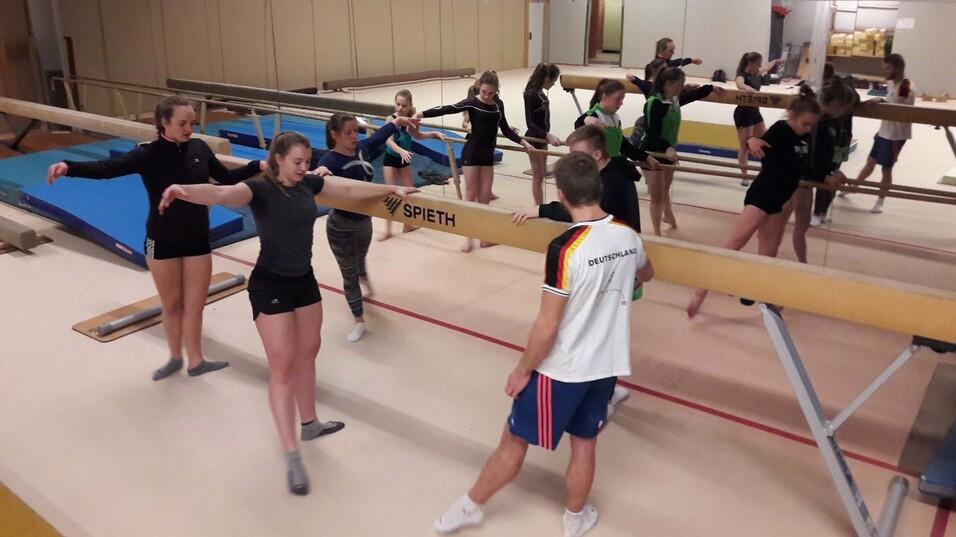 Gruppentraining von weiblichen Turnerinnen an einem Schwebebalken in einer Gymnastikhalle, mehrere Übungspaarungen.