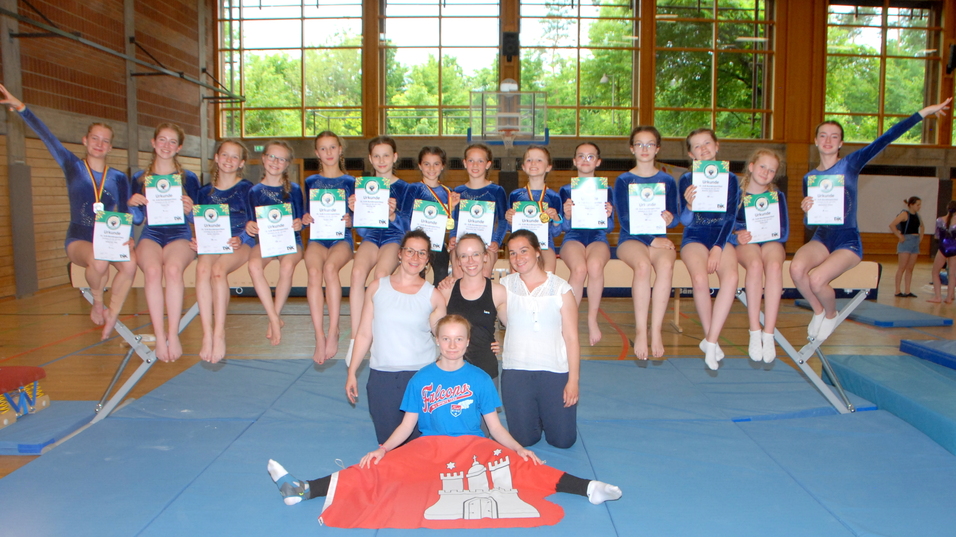 Gruppenfoto von 14 Turnerinnen mit Medaillen und Urkunden auf einem Sportger&auml;t in einer Turnhalle, umgeben von Trainerinnen.