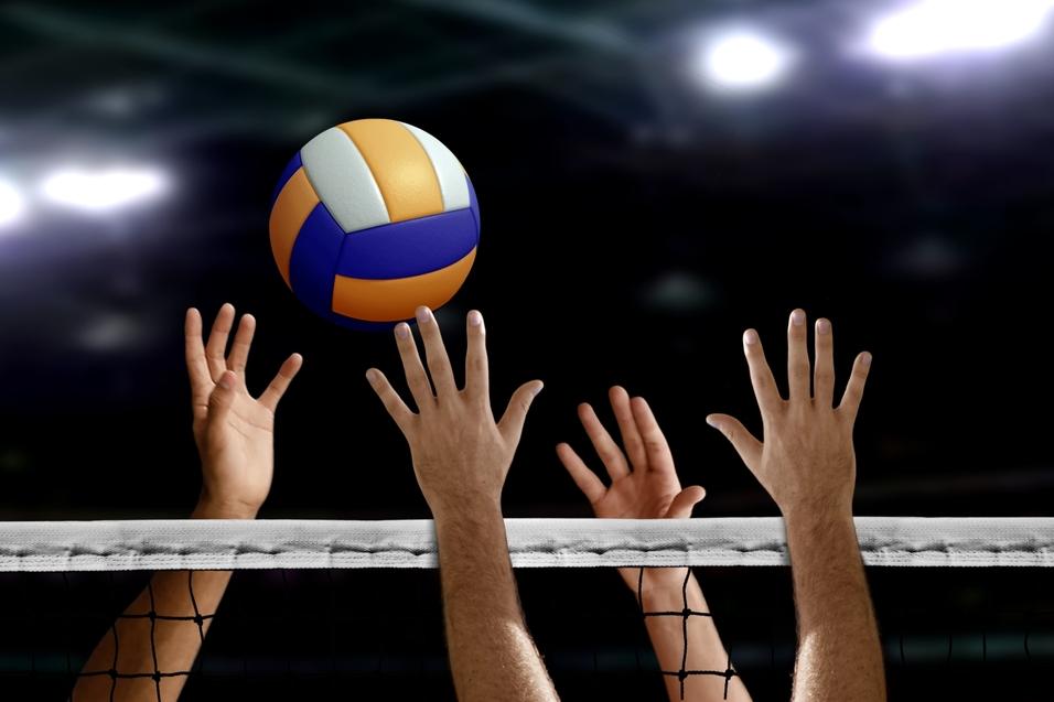 H&auml;nde greifen nach einem Volleyball &uuml;ber einem Netz in einer Sporthalle mit verschwommenem Hintergrund.