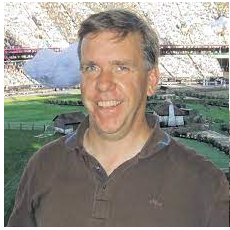 Ein Mann mit kurzem, braunem Haar steht l&auml;chelnd vor einer Sportarena mit einem gr&uuml;nen Feld im Hintergrund.