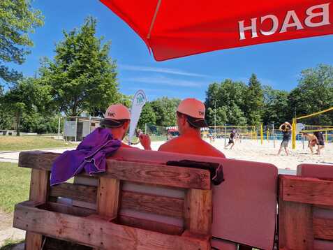 Zwei Personen mit wei&szlig;en Caps sitzen auf einer Holzbank und beobachten ein Volleyballspiel an einem Sandplatz.