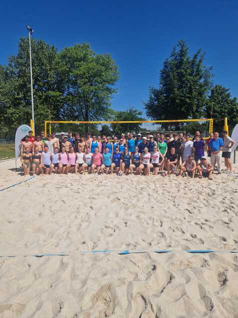 Gruppe von etwa 50 Personen vor einem Beachvolleyball-Netz auf einem Sandplatz, umgeben von B&auml;umen und blauem Himmel.