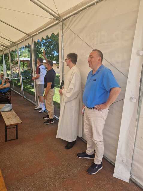 Gruppe von M&auml;nnern in einem Zelt, einer tr&auml;gt einen wei&szlig;en Habit, andere lehnen an der Wand. Tische und St&uuml;hle im Hintergrund.