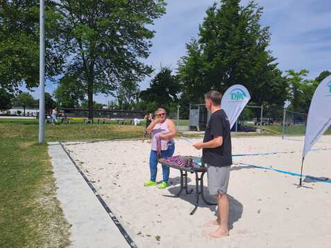 Zwei Personen stehen auf einem Sandplatz, eine spricht ins Mikrofon, w&auml;hrend ein Tisch neben ihnen steht.