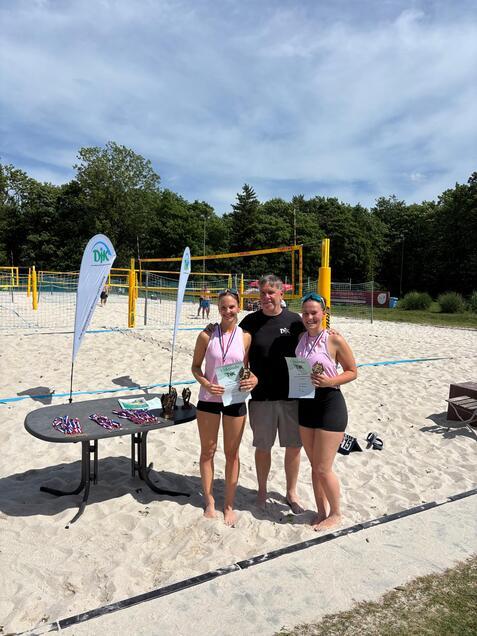 Zwei Frauen stehen mit Auszeichnungen und Urkunden neben einem Mann auf einem Sandvolleyballfeld.