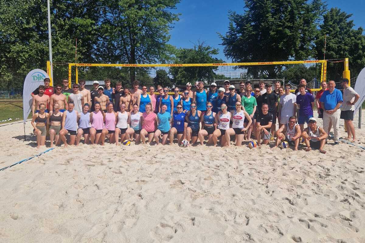 Gruppe von etwa 50 Personen vor einem Beachvolleyball-Netz auf einem Sandplatz, umgeben von B&auml;umen und blauem Himmel.