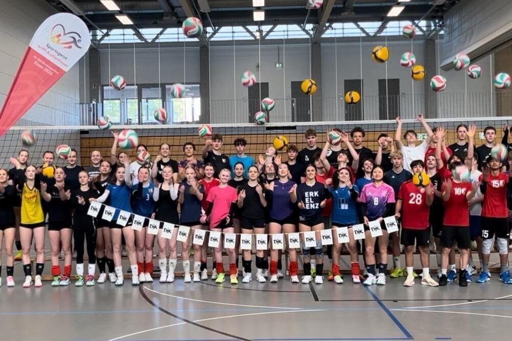 Gruppenshot von Volleyballspielern in der Halle, die B&auml;lle in die Luft werfen, umringt von Banner und Volleyballnetz.