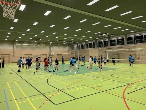 Vielzahl von Personen trainiert in einer Sporthalle im Volleyball, umgeben von Netzen und Basketballkorb.
