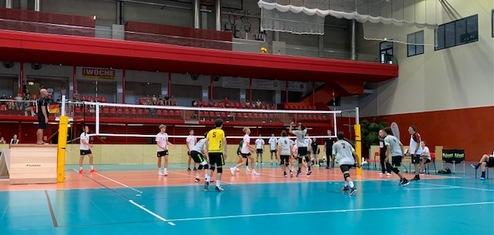 Volleyballspiel in einer Halle mit Spielern und Zuschauern, ein Spieler springt zum Setzen des Balls.