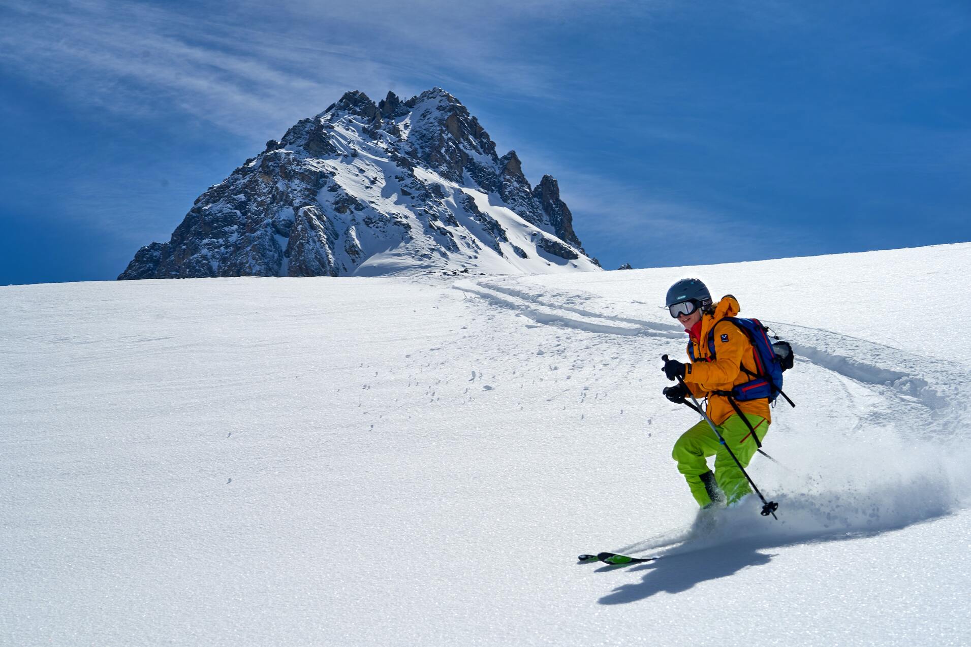 Skifahrer in leuchtend gelber Ausr&uuml;stung f&auml;hrt durch frischen Schnee mit einem schneebedeckten Berg im Hintergrund.