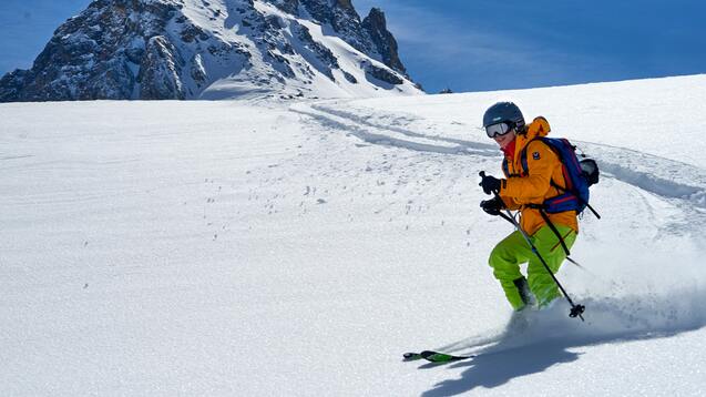 Skifahrer in leuchtend gelber Ausr&uuml;stung f&auml;hrt durch frischen Schnee mit einem schneebedeckten Berg im Hintergrund.