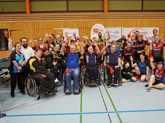 Gruppenszene von Tischtennisspielern, einschlie&szlig;lich Rollstuhlfahrern, die fr&ouml;hlich die H&auml;nde heben, in einer Sporthalle.