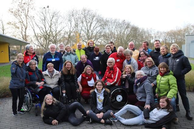 Gruppenzusammenkunft mit zahlreichen Menschen, darunter Rollstuhlnutzer, vor einer modernen Geb&auml;udeoberfl&auml;che im Freien.