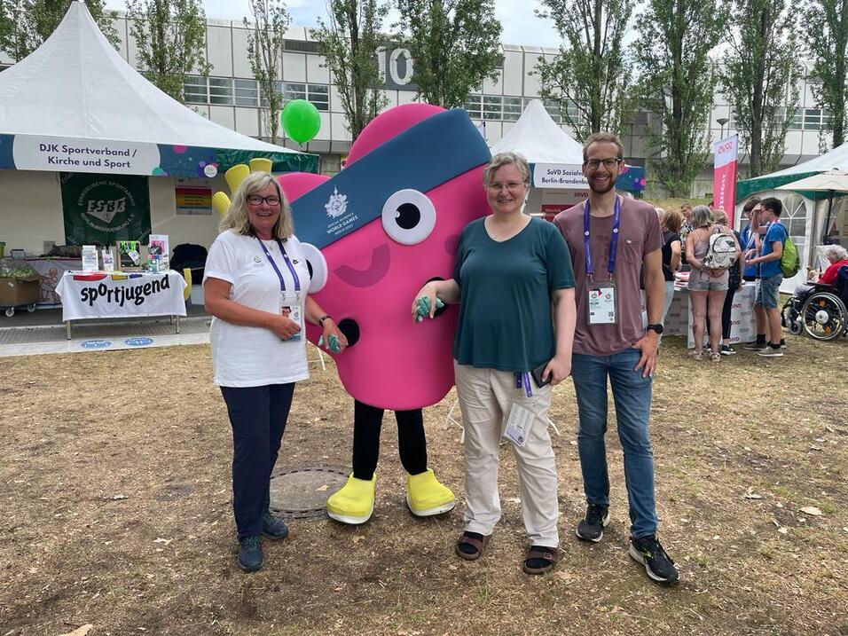 Drei Personen und ein pinkfarbener Maskottchencharakter stehen vor Zelten bei einer Veranstaltung im Freien.