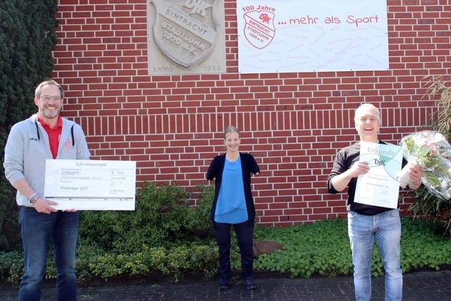 Drei Personen stehen vor einer Ziegelwand, halten einen Scheck, eine Urkunde und einen Blumenstrau&szlig;.