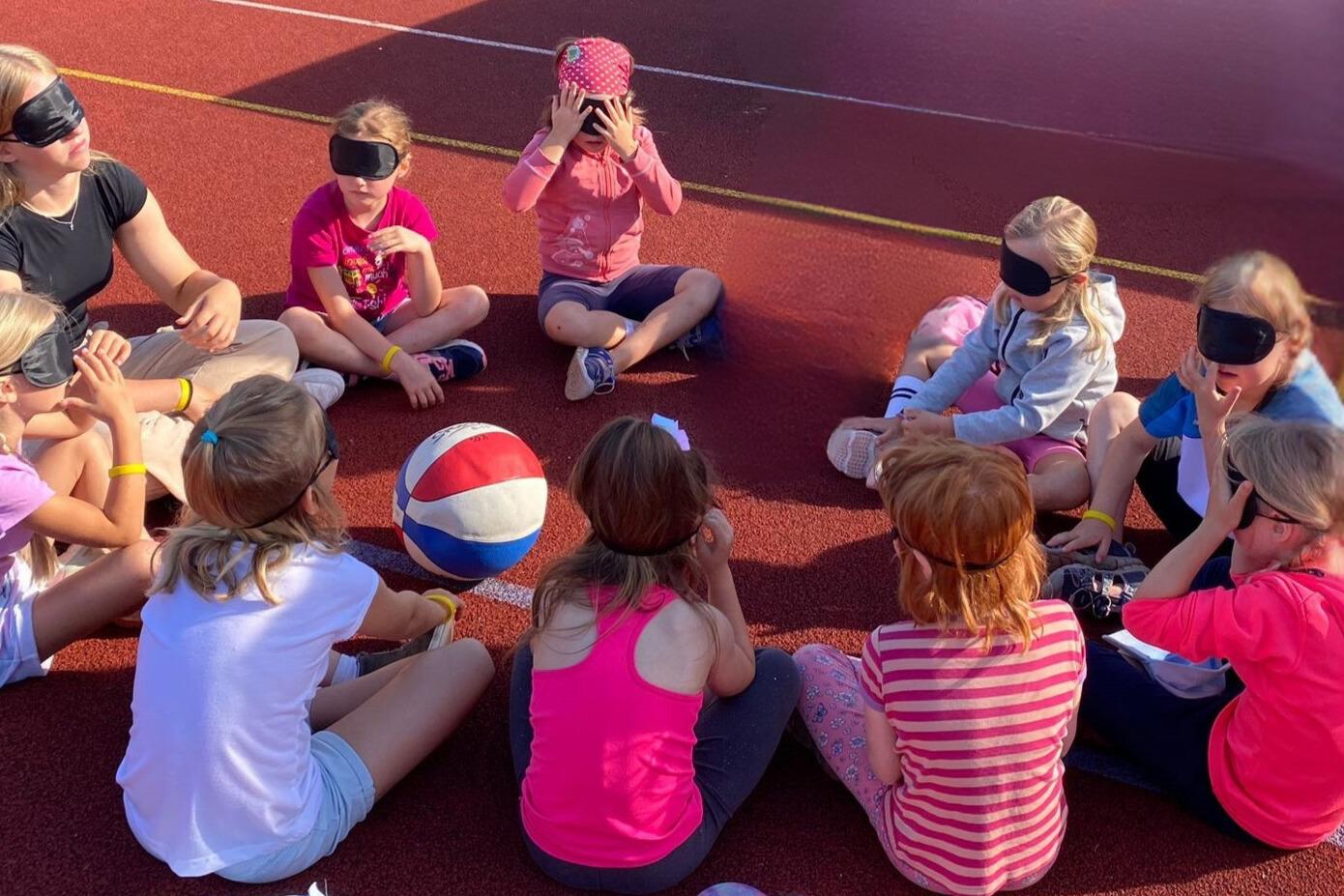 Gruppe von Kindern mit Augenbinden, die im Kreis auf einem Sportplatz sitzen. Ein Ball liegt in der Mitte.