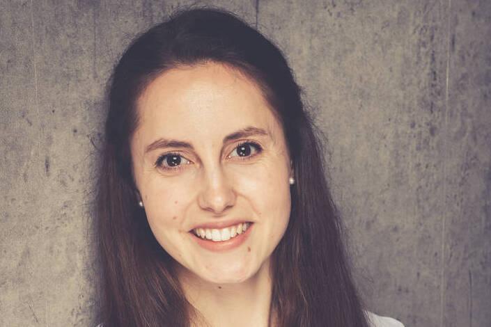 Junge Frau mit langen, braunen Haaren, l&auml;chelt in einem hellblauen Hemd vor einer grauen Betonwand.