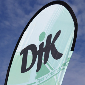 Fahne des DJK Sportverbands mit gr&uuml;nem Hintergrund und schwarzem Logo vor einem blauen Himmel.