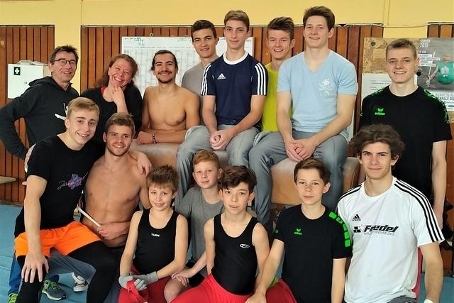 Gruppe von Jugendlichen und Trainern l&auml;chelt auf einem Gymnastikboden, umgeben von Sportger&auml;ten und einer Wandtafel.