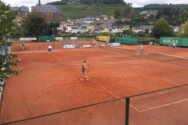 Tennisplatz mit Spielern auf roter Asche, umgeben von typischer Architektur und Weinbergen im Hintergrund.